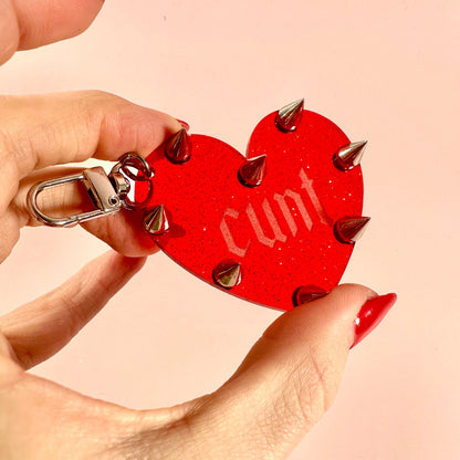 Red heart-shaped keychain with spikes held by a hand against a beige background