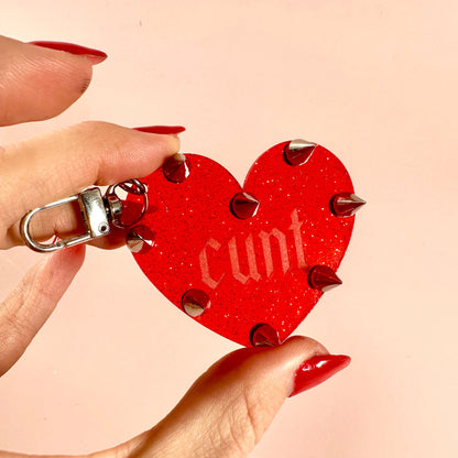 Red heart-shaped keychain with spikes held by a hand against a beige background