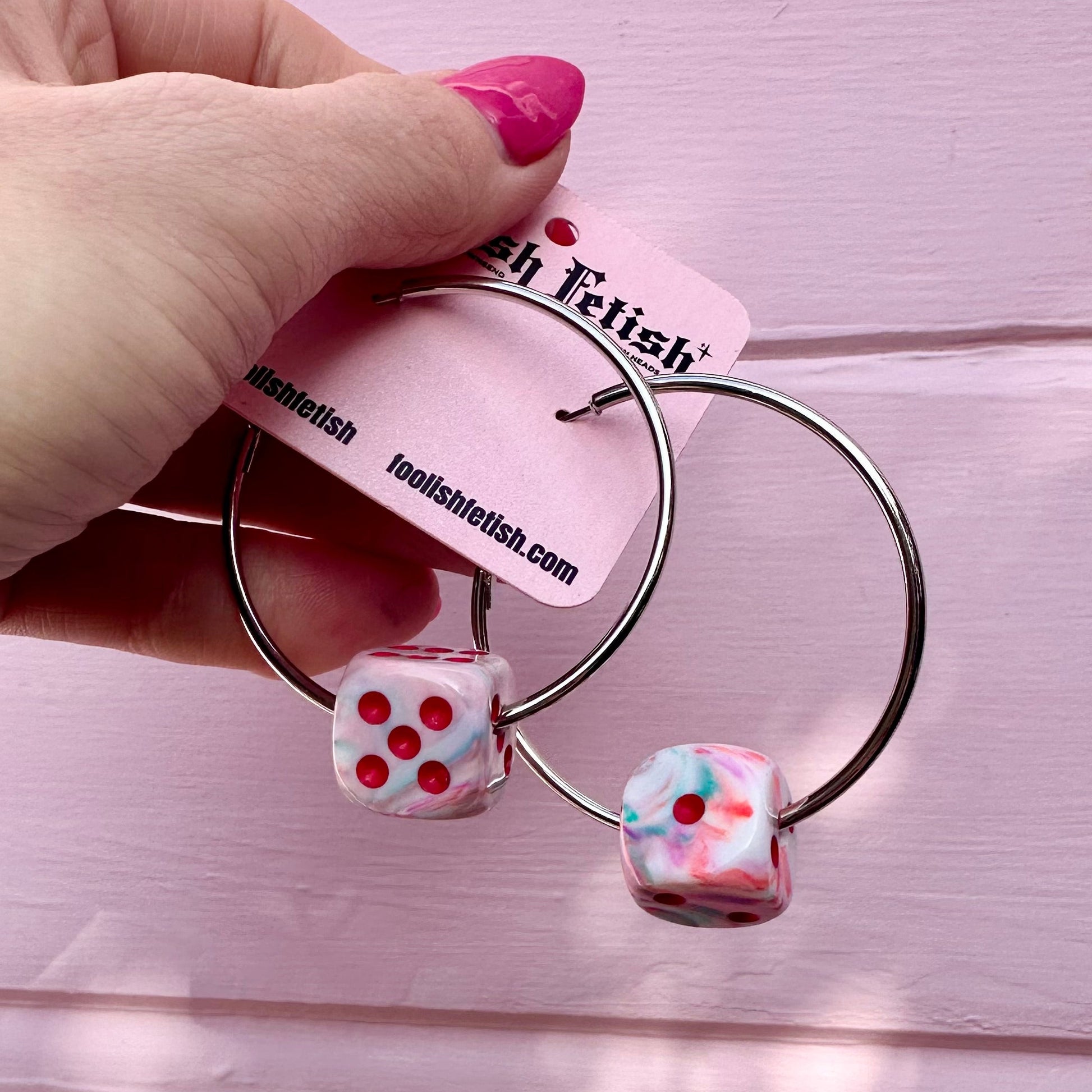 Earrings with dice-shaped charms held by a hand against a pink background