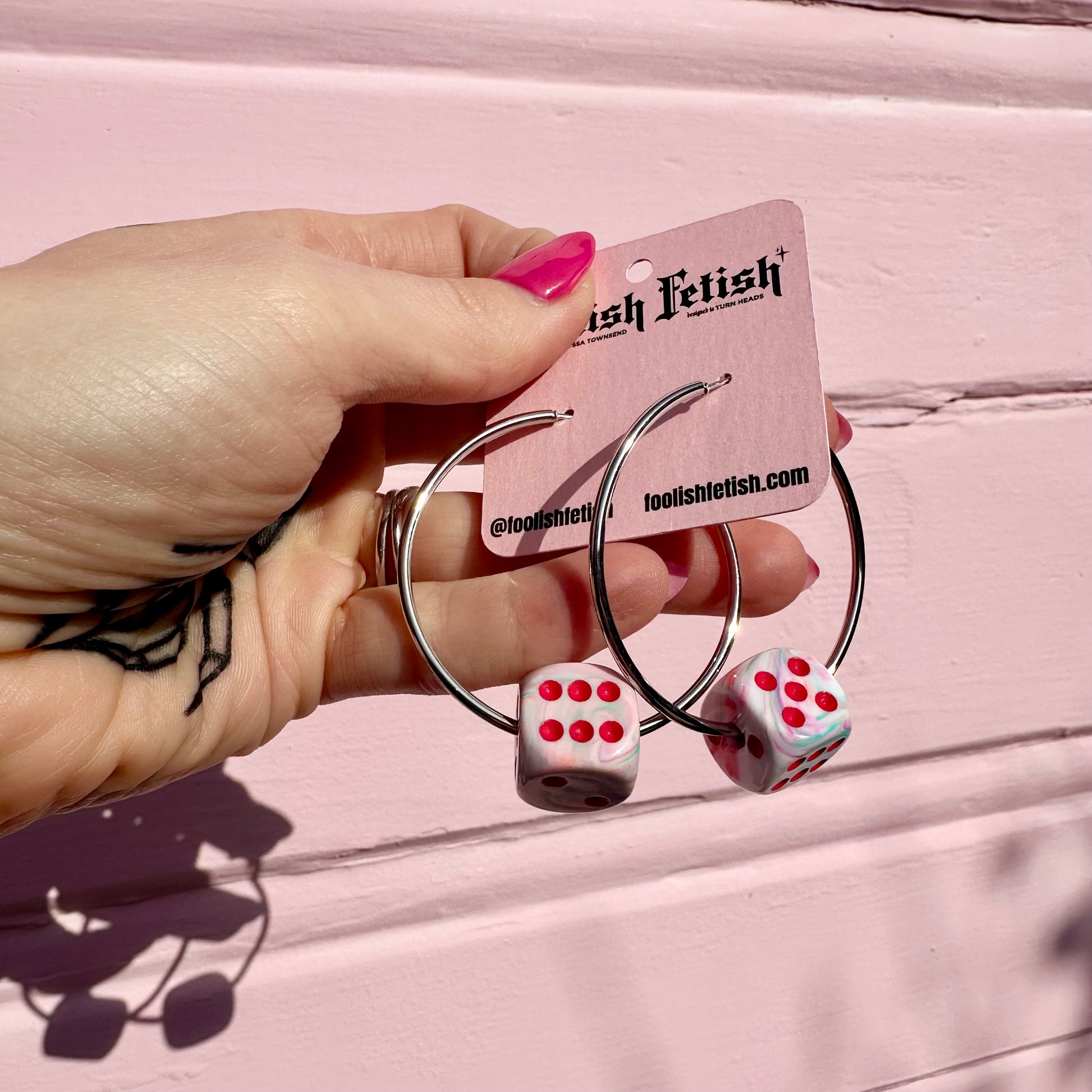 Hoop earrings with dice-shaped charms held by a hand against a pink background