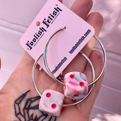 Hoop earrings with dice-shaped charms held by a hand, branded 'Foolish Lush'.