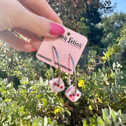 Dice-shaped earrings on a pink card held in front of green foliage