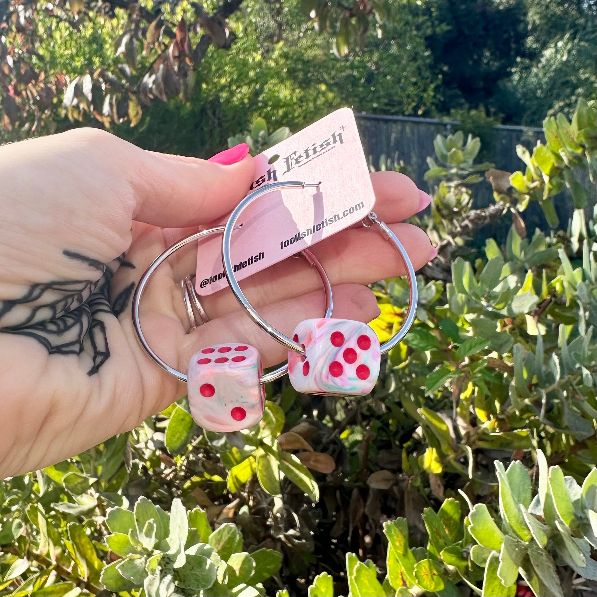 Hand holding pink dice-shaped earrings with a blurred green outdoor background