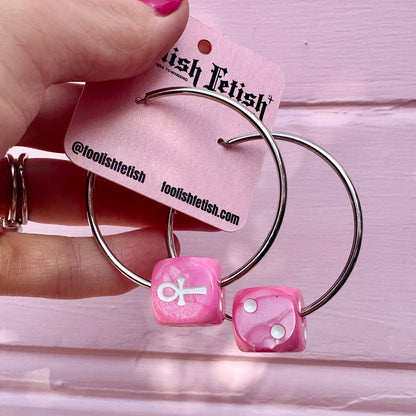 Pink dice-shaped earrings with metal hoop design held by a hand against a pink background.