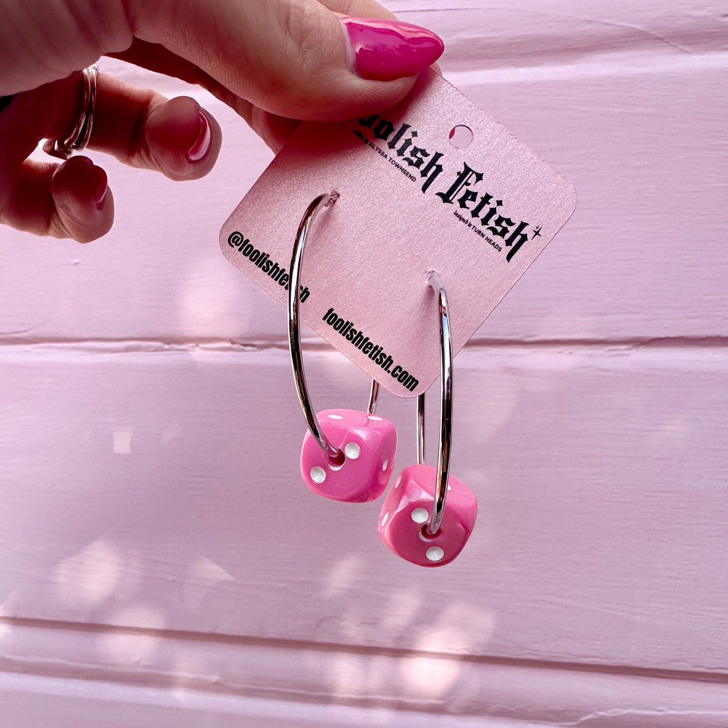 Pink dice-shaped earrings on a 'Foolish Fetish' branded card against a pink background.