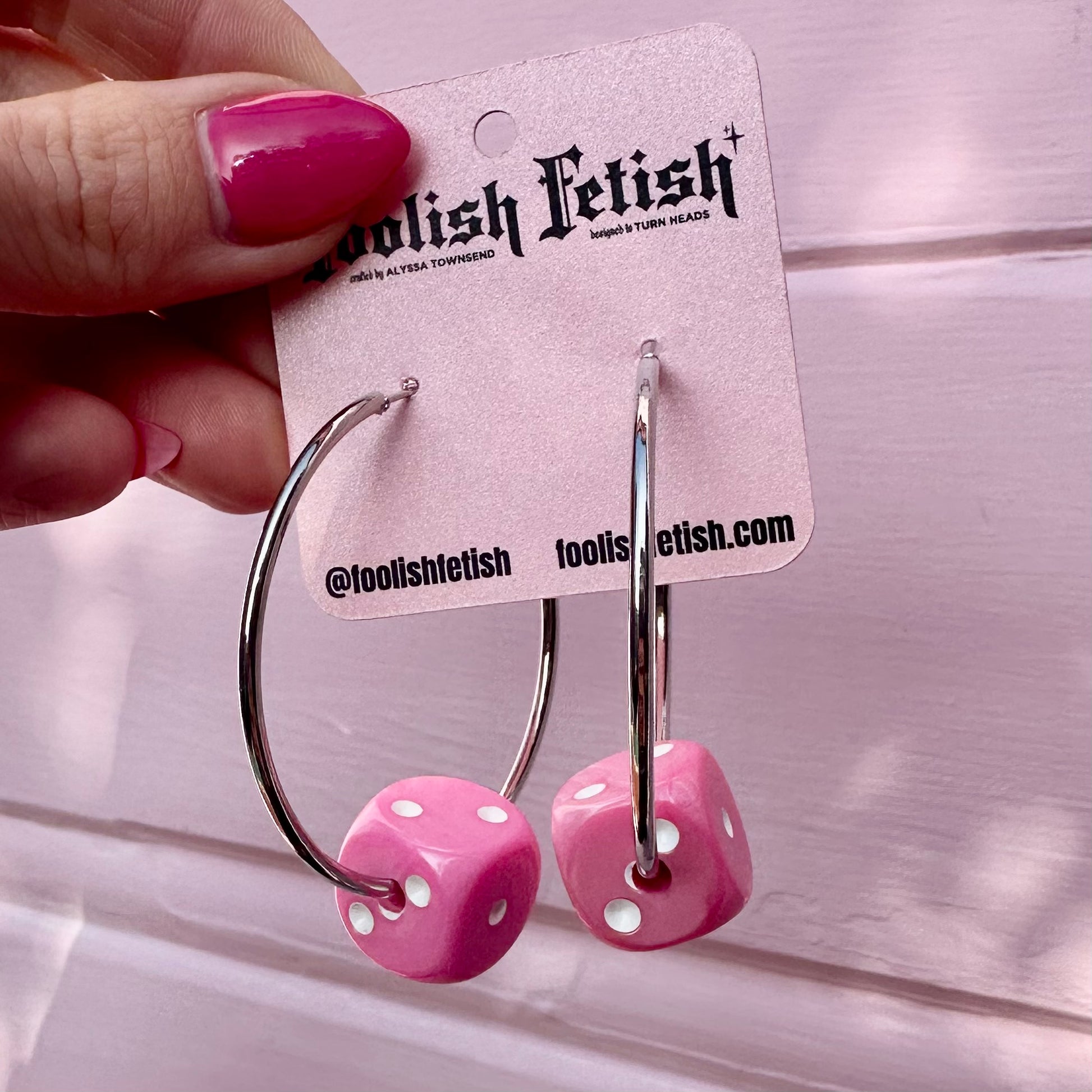 Pink dice-shaped earrings on a 'Foolish Fetish' card held by a hand with pink nail polish.