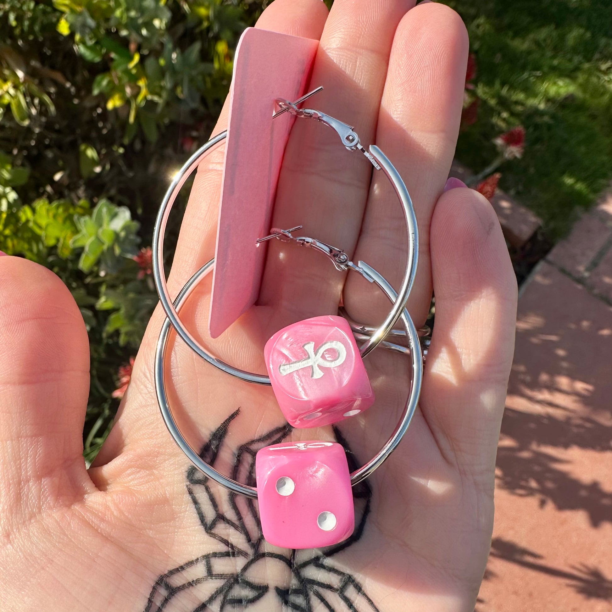 Hand holding pink dice with a symbol and silver hoop earrings against a natural background