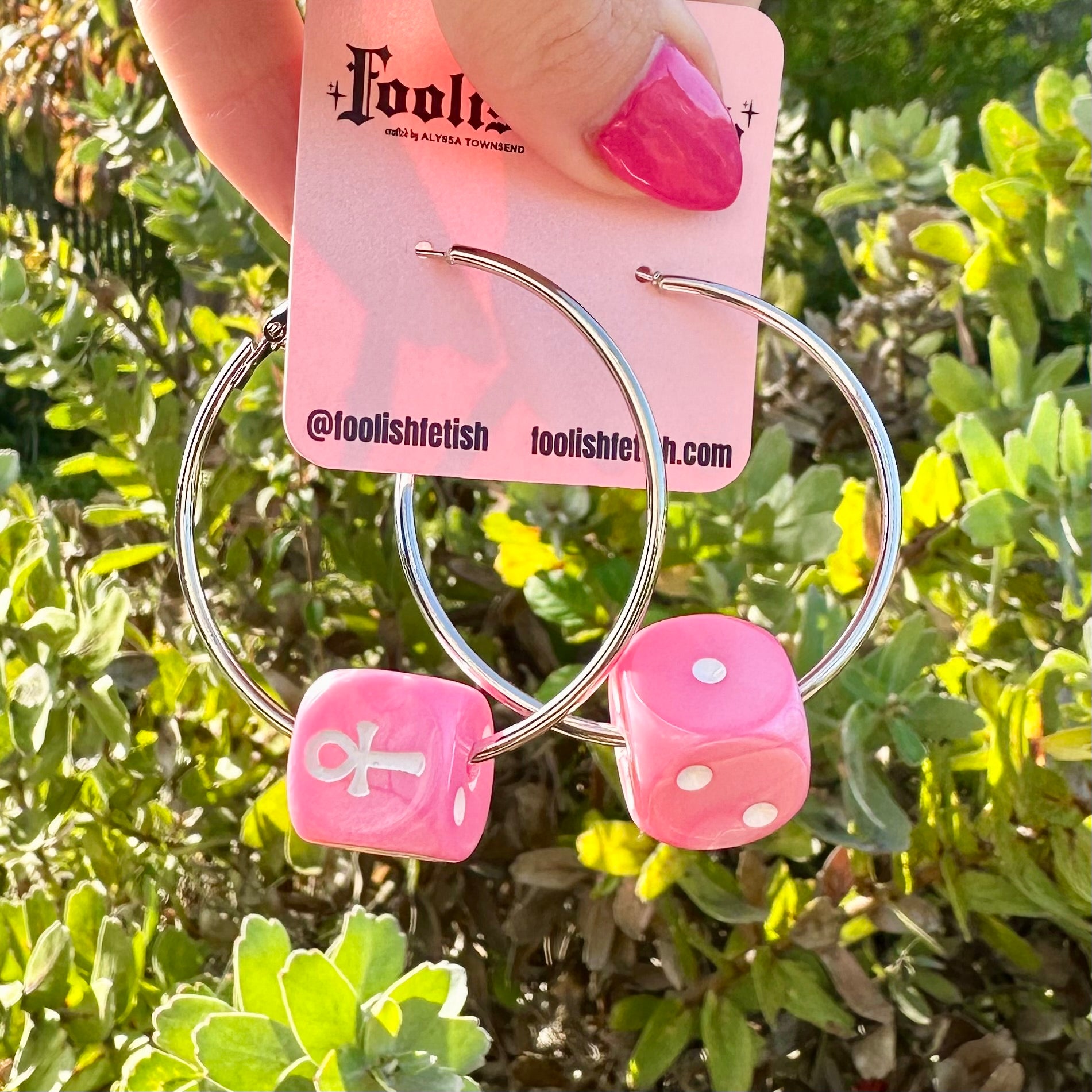 Pink dice-shaped earrings with silver hoop design on a green leafy background