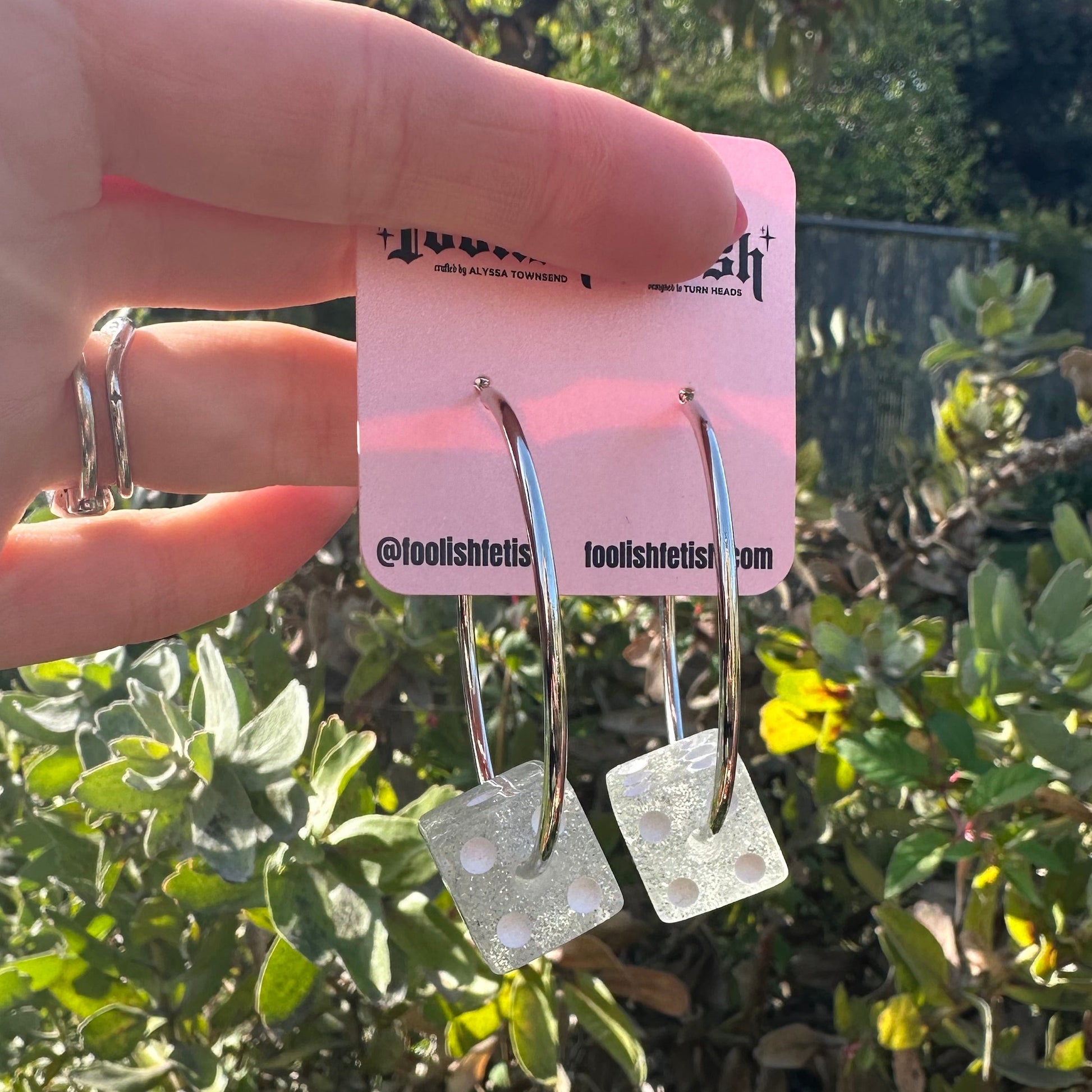 Hand holding a pair of earrings with a pink card against a green leafy background
