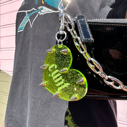Green glittery heart-shaped charm with 'Cunt' on a black bag.