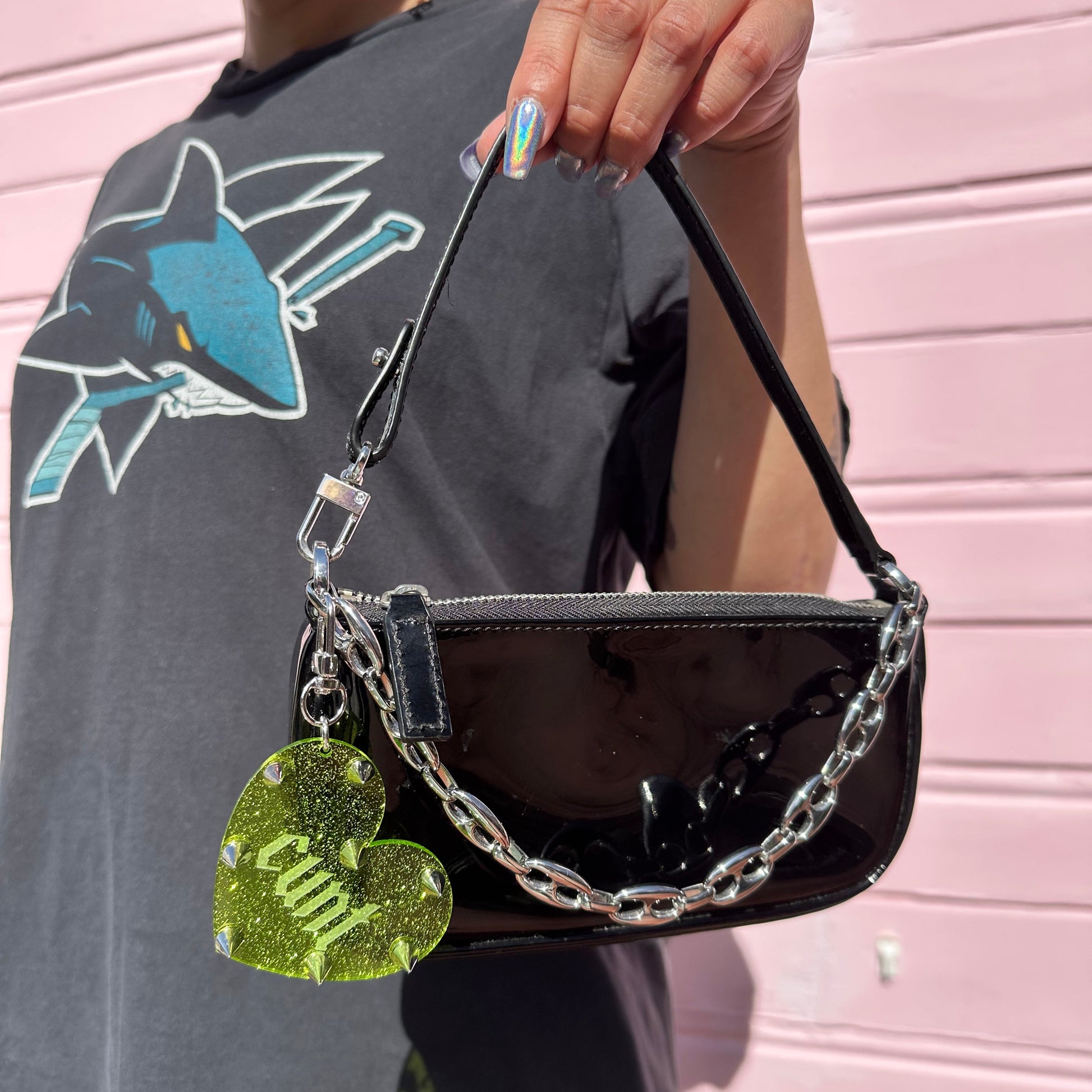 Person holding a black handbag with a chain detail and a green heart charm against a pink background
