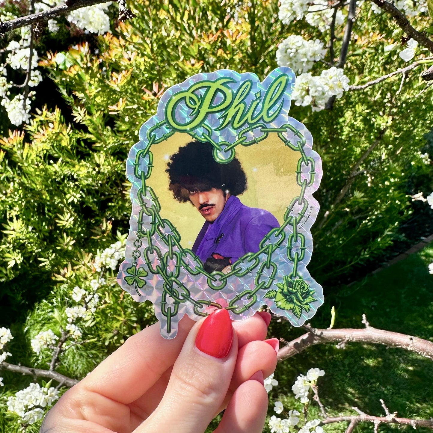 Hand holding a sticker with a portrait and 'Phil' text against a floral background