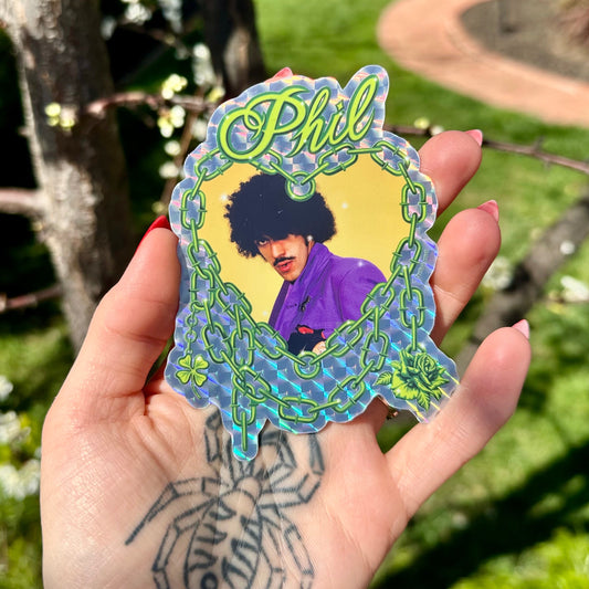 Hand holding a 'Phil' sticker with a colorful frame against a garden background