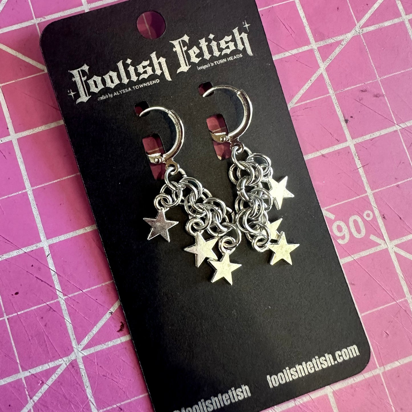 Silver chain and star earrings on a Foolish Fetish branded card against a pink grid background.
