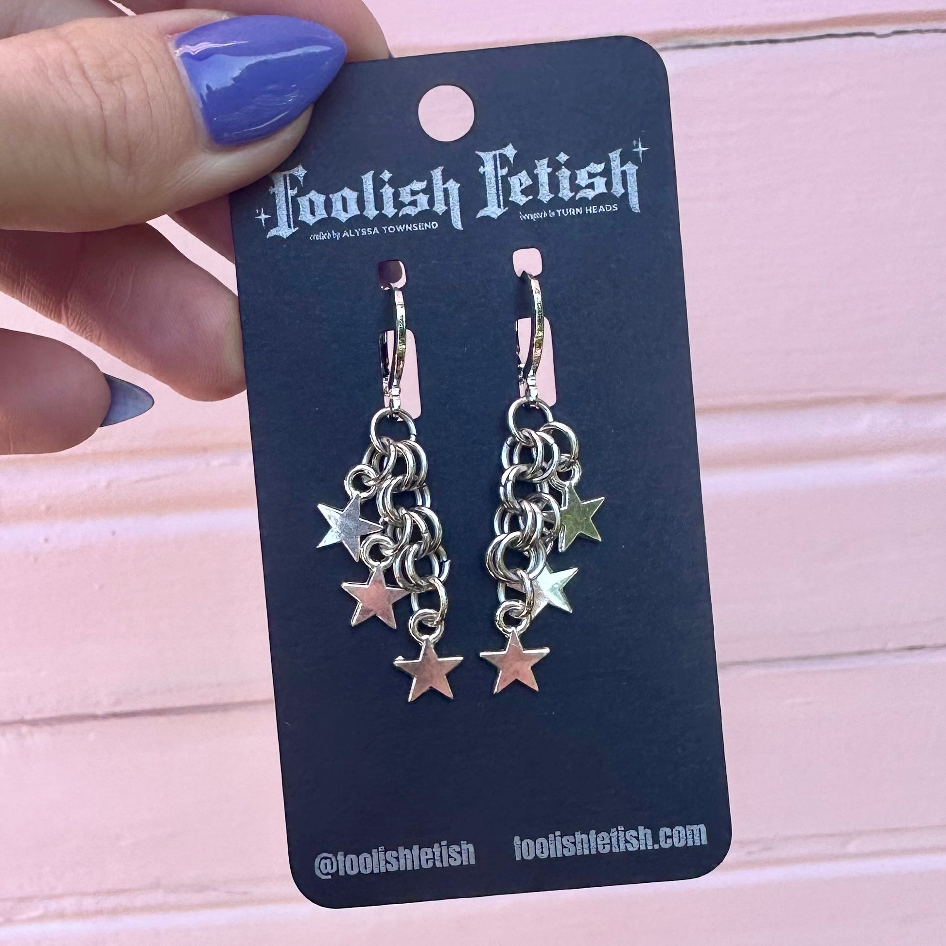 Star-shaped earrings on a Foolish Fetish branded card held by a hand with purple nail polish.