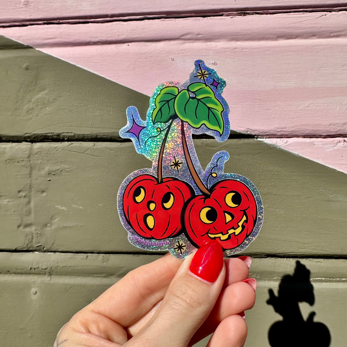 A hand with red nail polish holds a die-cut holographic sticker of two smiling jack-o'-lantern cherries against a background of pink and olive green painted wood planks.
