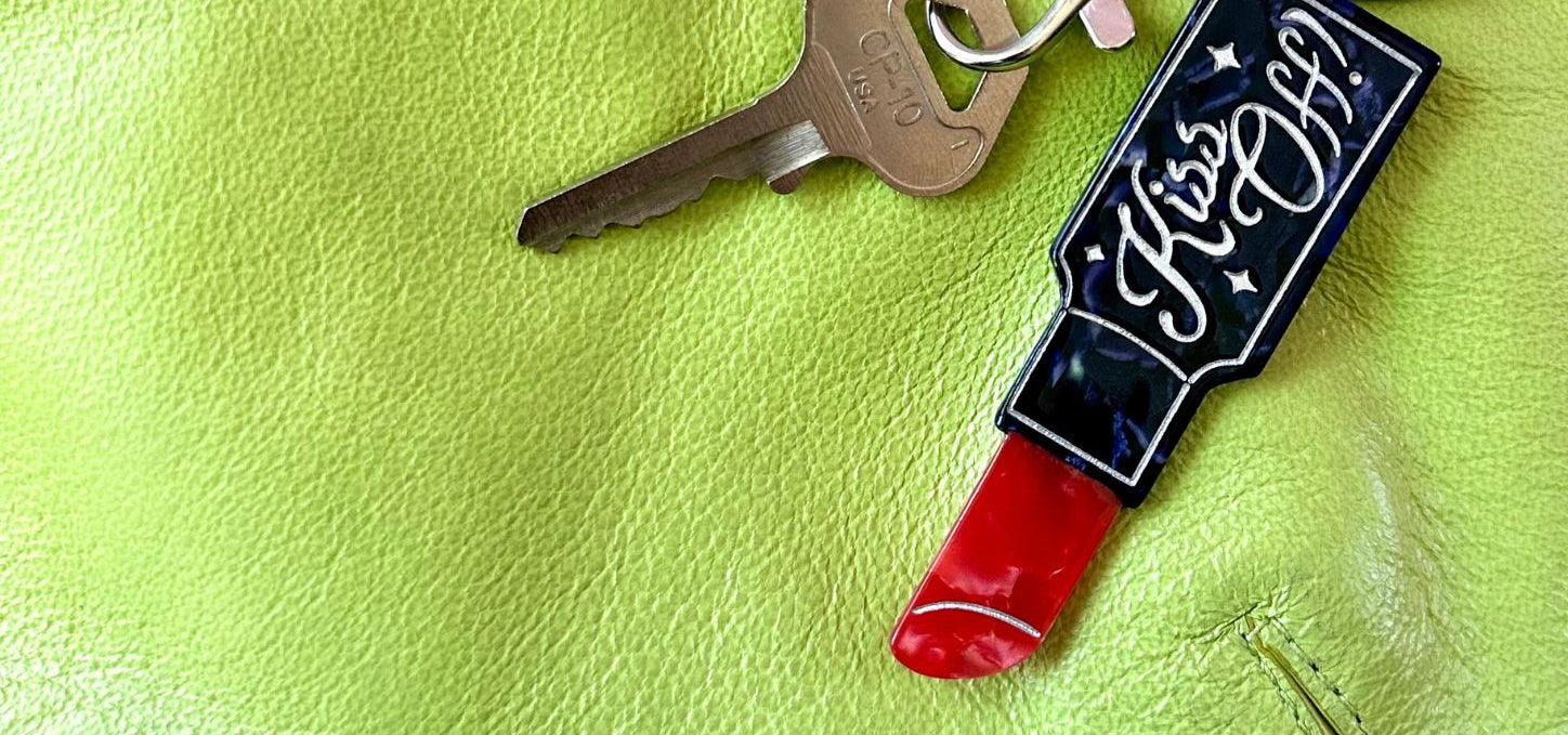 A close-up of a keychain with a lipstick-shaped charm and a key on a bright lime green textured surface against a pink background.