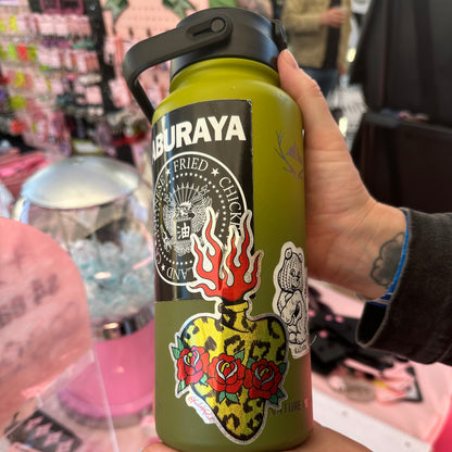 A person holds a green insulated water bottle with a black lid and multiple colorful stickers, including a skull, flames, and roses. The background is a busy retail display with pink items.