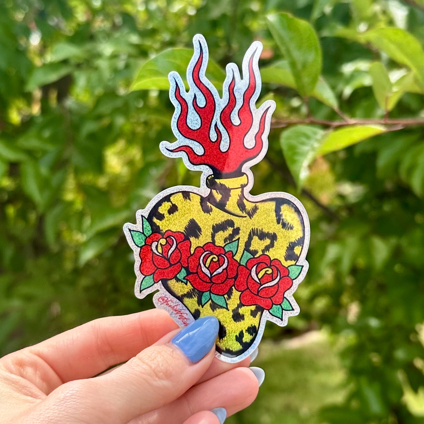 A hand with blue nail polish holds a die-cut sticker featuring a traditional tattoo-style heart with red roses, a leopard print background, and red flames at the top, set against a blurred green garden background.