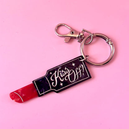 A lipstick-shaped keychain with a red lipstick tip, a black body, and the words 'Kiss Off!' in white script, attached to a silver metal split ring and clasp, set against a solid pink background.