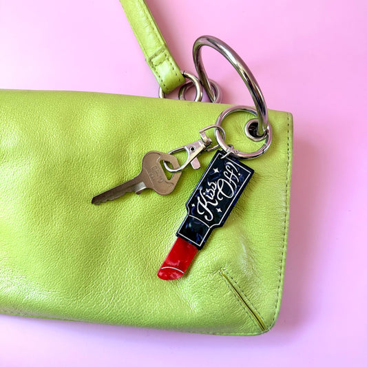 A close-up of a lime green leather wristlet clutch with a silver metal ring and keychain attachment. The keychain features a black enamel lipstick-shaped charm with the words 'Kiss My Lips' in white script, alongside a silver key.