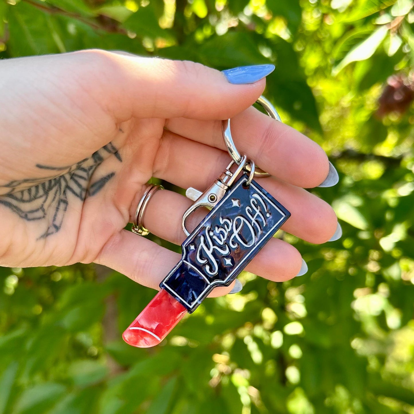 A hand holding a lipstick-shaped keychain outdoors in front of green leaves. The keychain has a dark blue lipstick barrel with white script reading 'Fate is her', a red lipstick tip, and a silver key ring.