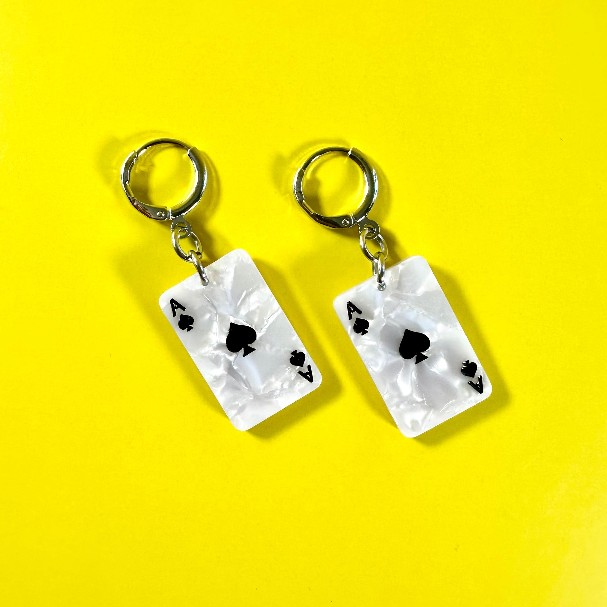 A pair of dangling earrings with rectangular white playing card charms featuring black aces of spades and four of clubs, suspended from silver hoop backings, set against a solid bright yellow background.