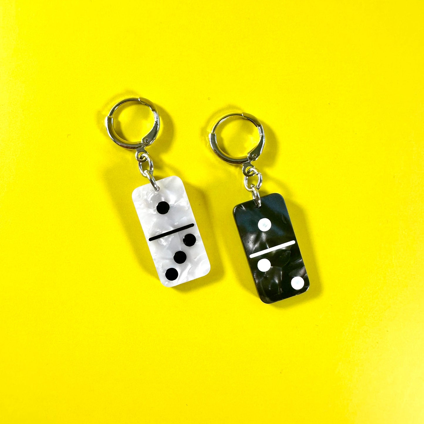 Two domino-shaped earrings on a bright yellow background. One earring has a white domino face with black pips and a silver earring, and the other has a black domino face with white pips and a silver key ring.
