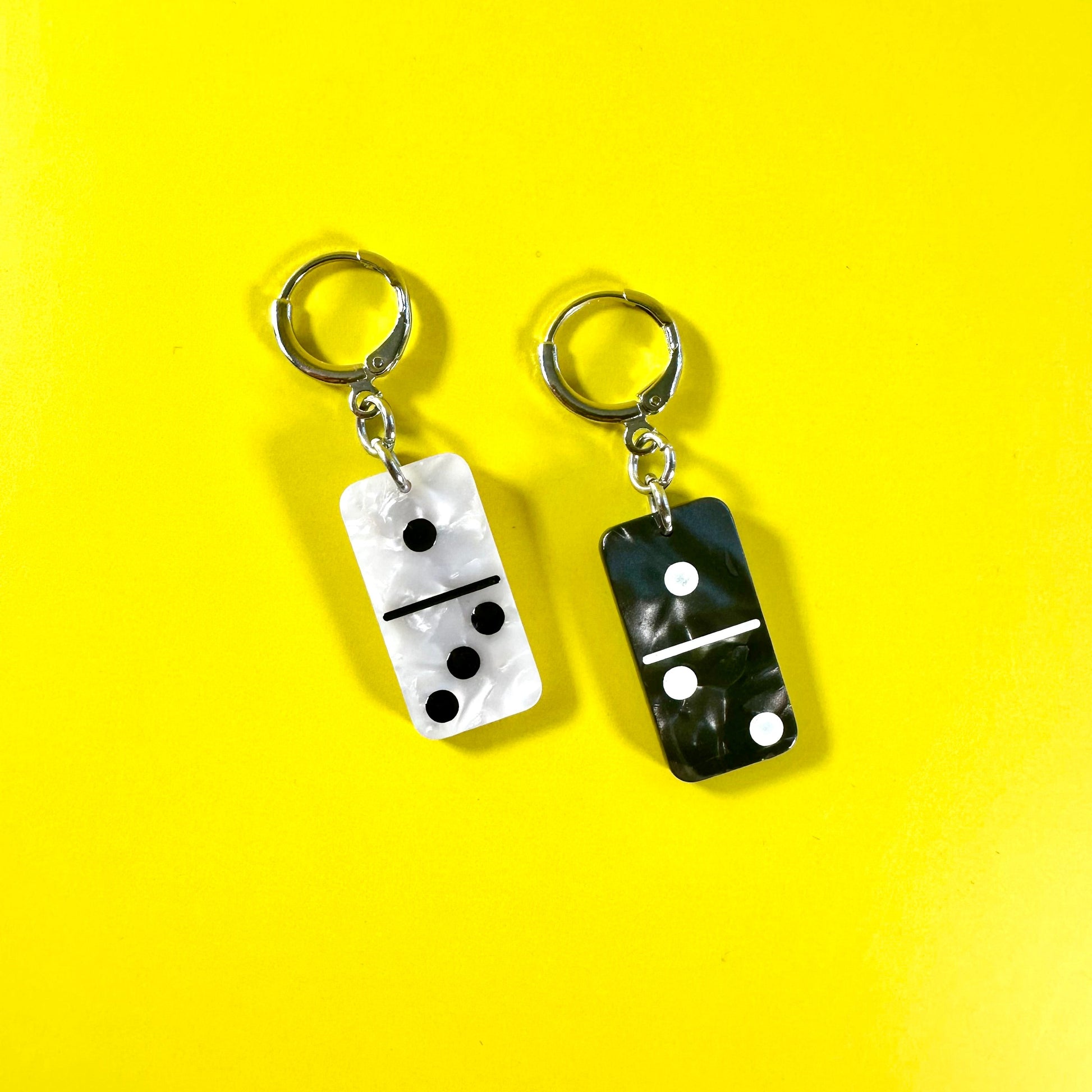 Two domino-shaped earrings on a bright yellow background. One earring has a white domino face with black pips and a silver earring, and the other has a black domino face with white pips and a silver key ring.