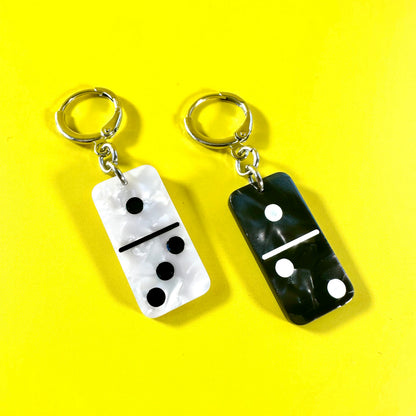 Two domino-shaped earrings on a bright yellow background. One earring has a white domino with black pips and a silver earring, and the other has a black domino with white pips and a silver key ring.