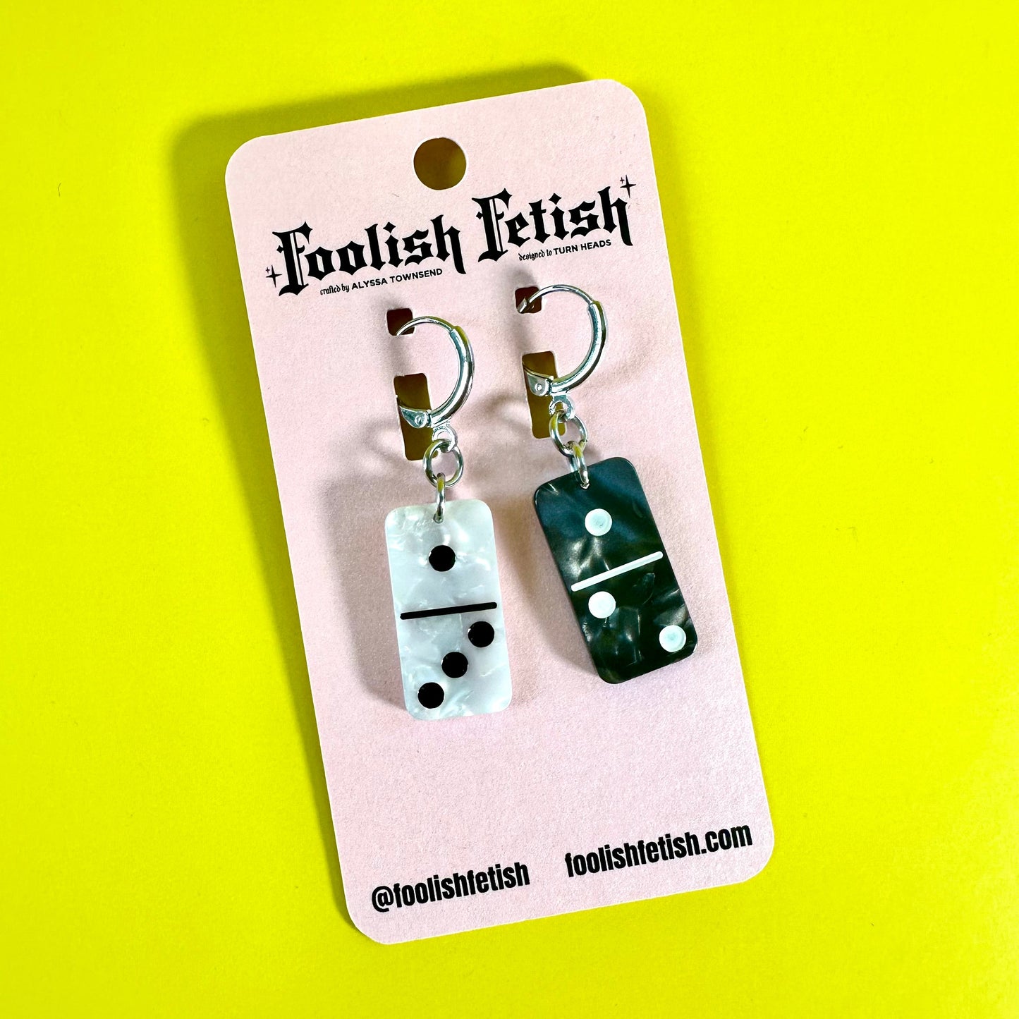 A pair of novelty dangle earrings displayed on a pink retail card. The earrings feature rectangular domino-shaped charms, one white with black pips and one dark green with white pips, attached to gold-toned hoops with small brown beads.