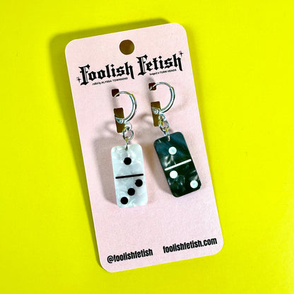 A pair of novelty dangle earrings displayed on a pink retail card. The earrings feature rectangular domino-shaped charms, one white with black pips and one dark green with white pips, attached to gold-toned hoops with small brown beads.