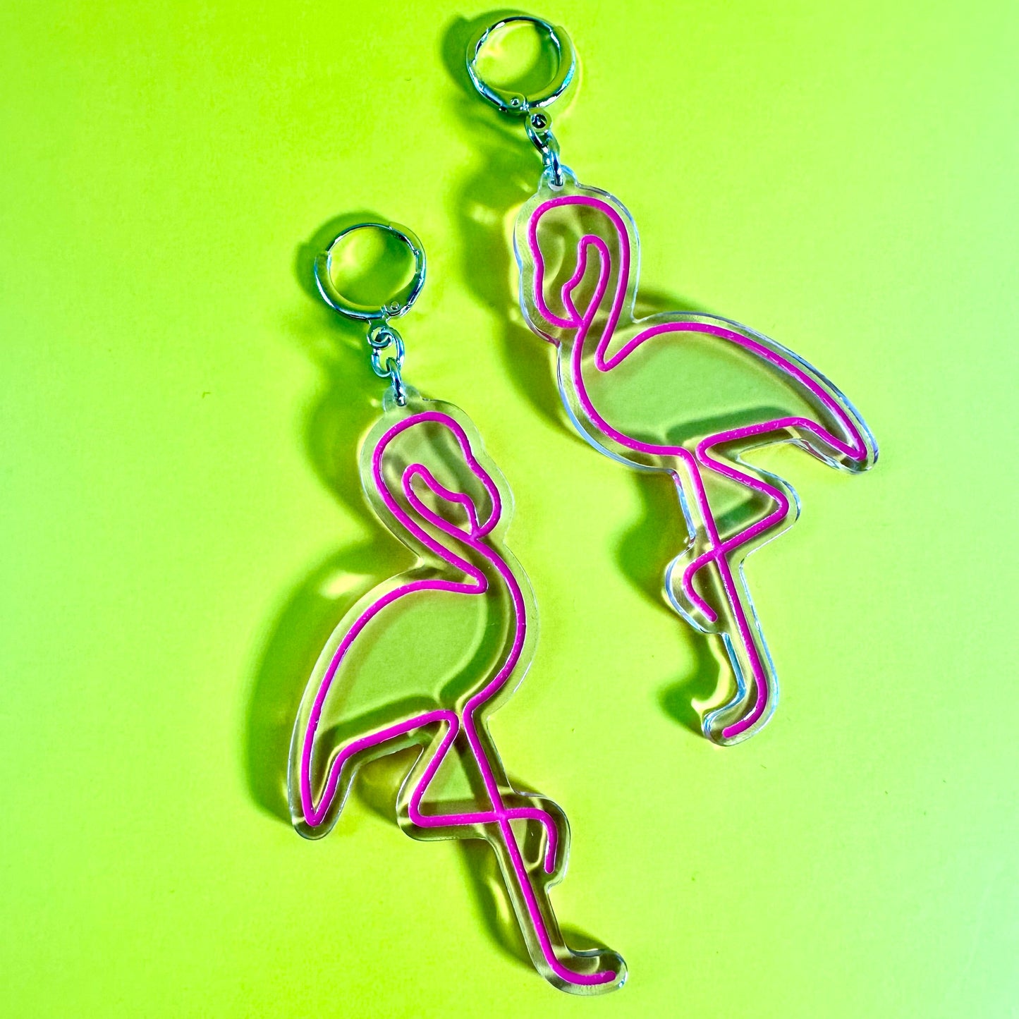 A pair of flamingo-shaped acrylic earrings  with silver-tone metal loops, displayed on a bright lime green background.