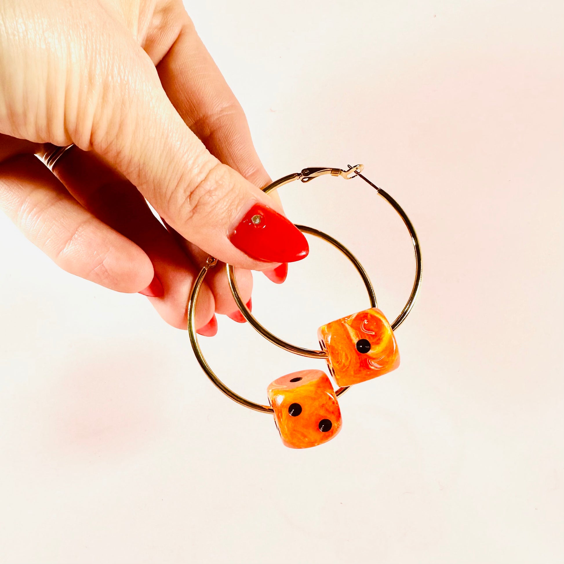 Hand holding gold hoop earrings with orange dice-shaped charms on a light pink background