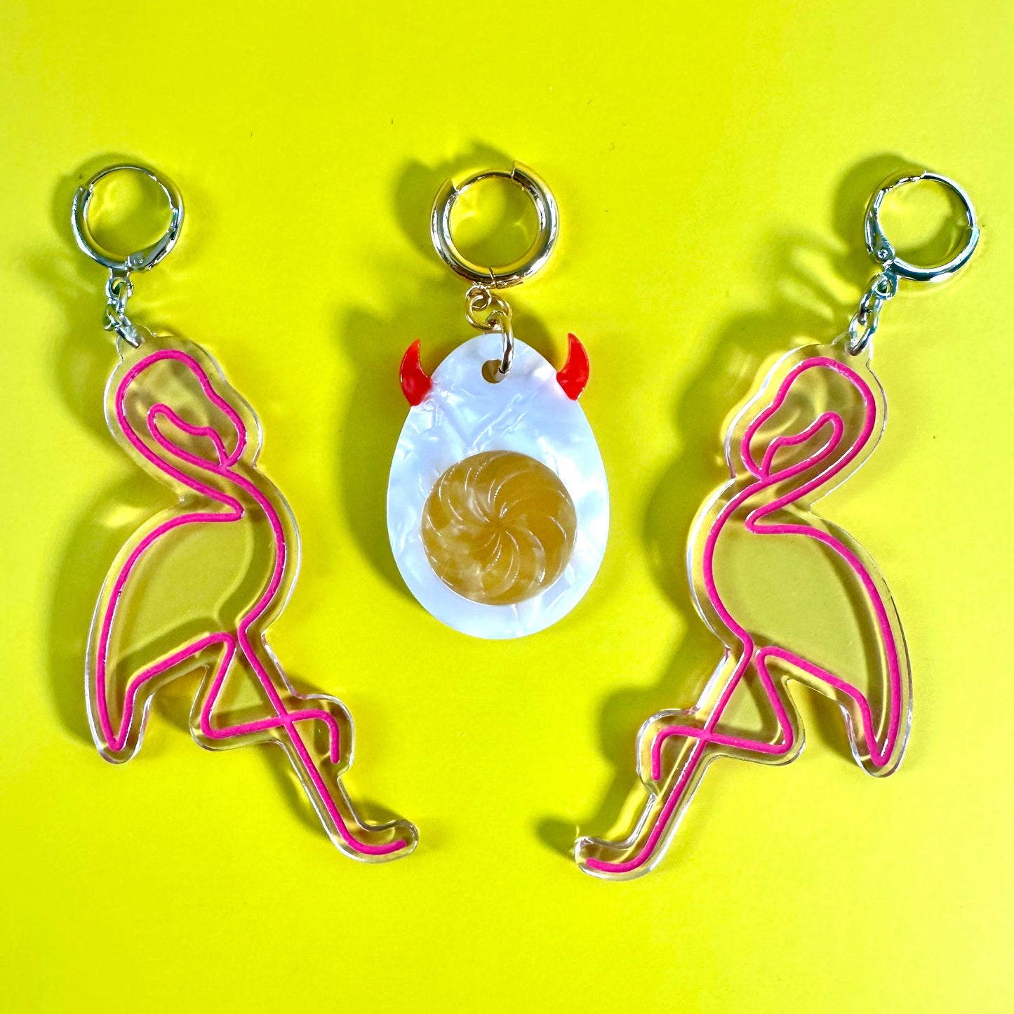 Three flamingo-themed earrings arranged on a bright yellow background. Two keychains feature pink flamingo outlines with silver-tone rings, and the center earring features a white egg pendant with small red horns and a gold swirl center.