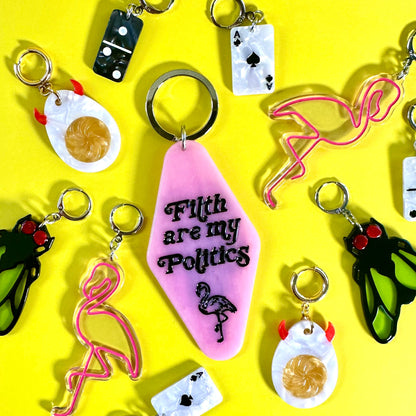 A collection of colorful novelty keychains arranged on a bright yellow background. The central keychain is pink with the text 'F*th are my Politics' and a flamingo illustration.