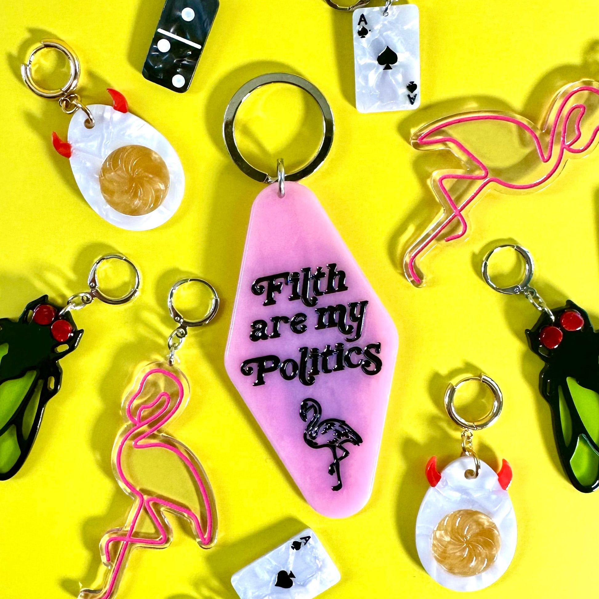 A collection of colorful novelty earrings arranged on a bright yellow background. The central keychain is pink with the text 'F*th are my Politics' and a flamingo illustration.