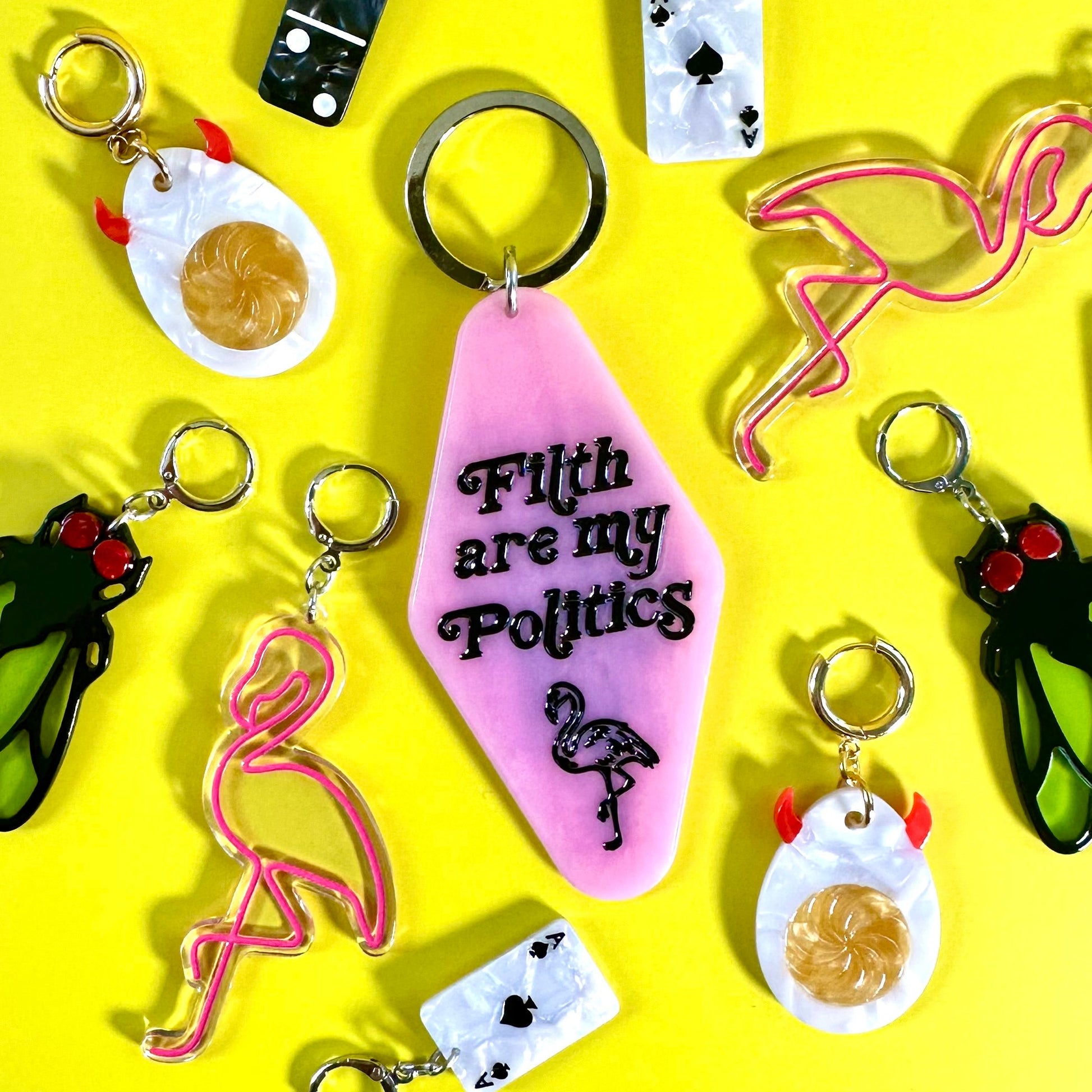 A collection of colorful novelty earrings arranged on a bright yellow background. The central keychain is pink with the text 'F*th are my Politics' and a flamingo illustration.