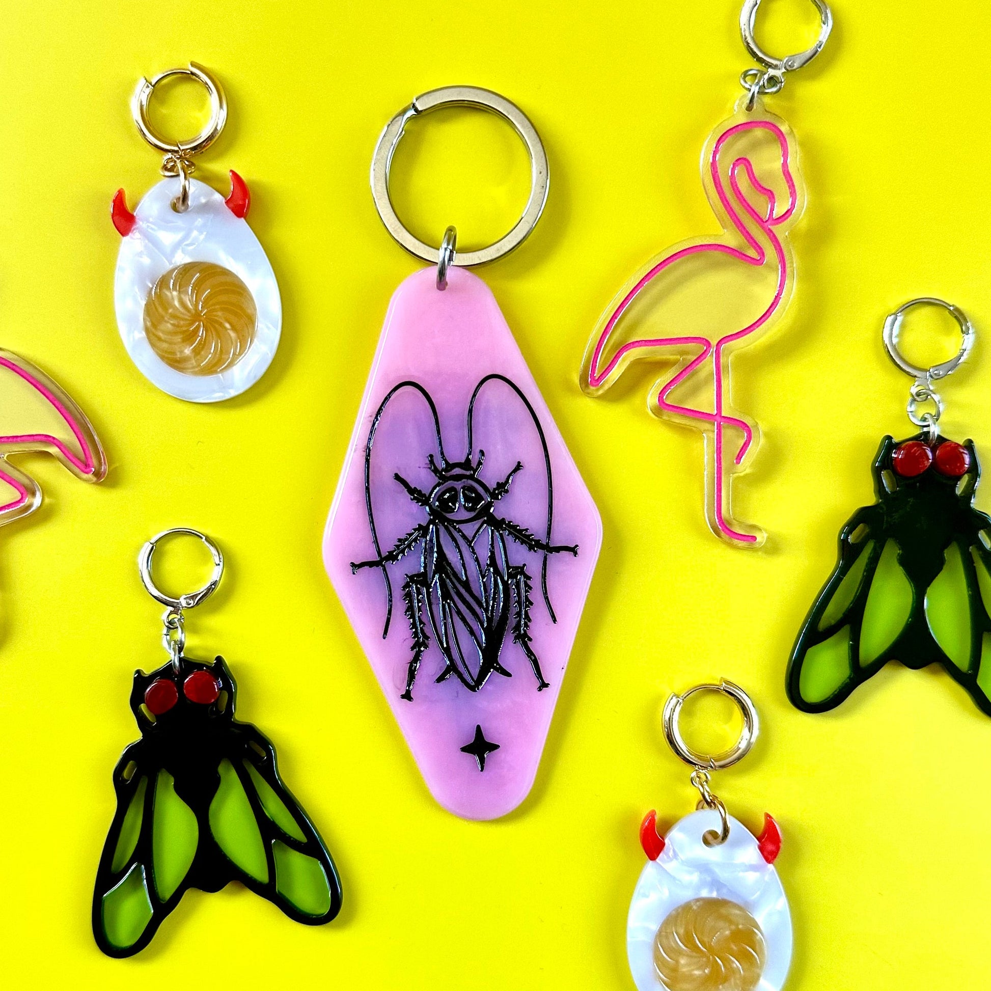 A set of six novelty keychains arranged on a bright yellow background. The collection includes flamingo and fly designs in pink, black, and green, as well as cracked-egg keychains in white and orange.