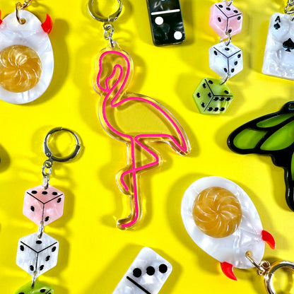 A collection of colorful acrylic earrings arranged on a bright yellow background. The earrings feature whimsical designs including pink flamingo, dice, eggs, a butterfly, and a small boat.