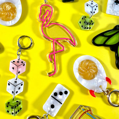 A collection of colorful acrylic earrings arranged on a bright yellow background. The earrings feature whimsical designs including pink flamingo, dice, eggs, a butterfly, and a small boat.