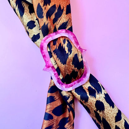 A leopard print scarf with brown, black, and beige tones, tied in a knot with a pink and white patterned ring against a solid lavender background.