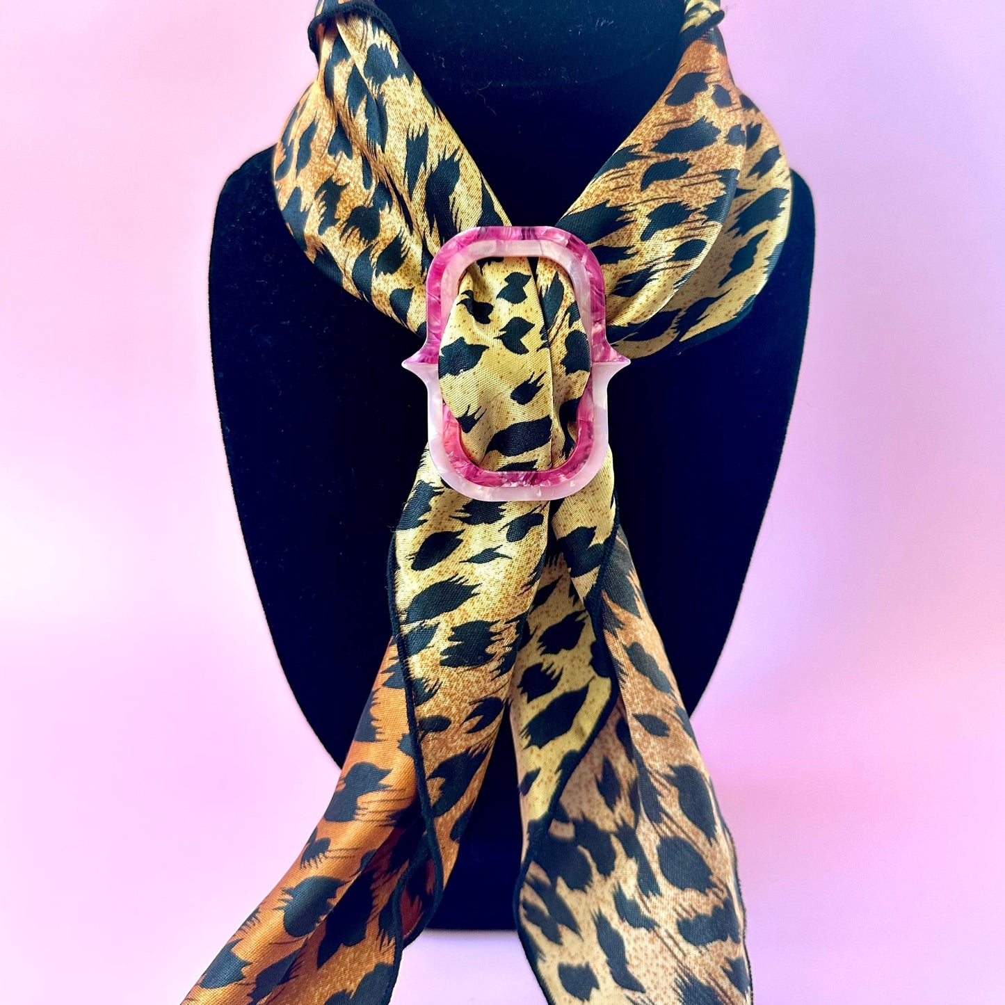 A leopard print scarf with a pink scarf buckle, displayed on a black mannequin bust against a pink background.