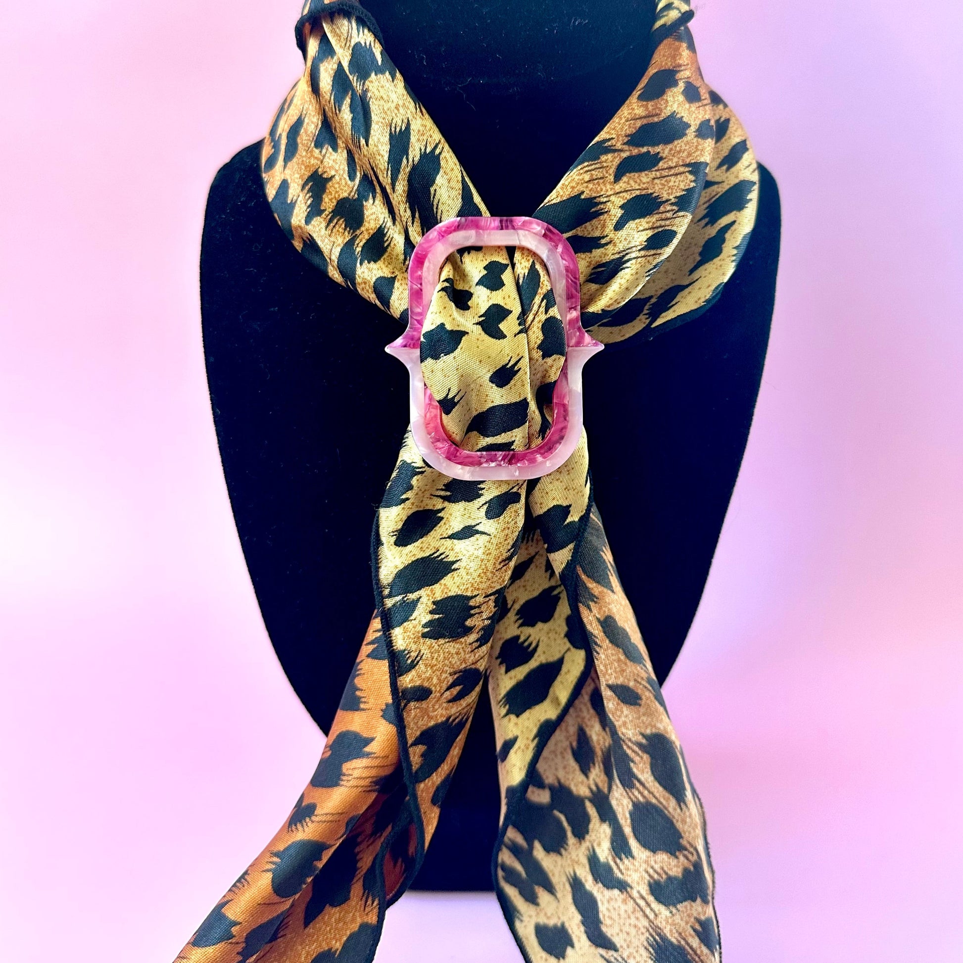 A leopard print scarf with a pink scarf buckle, displayed on a black mannequin bust against a pink background.