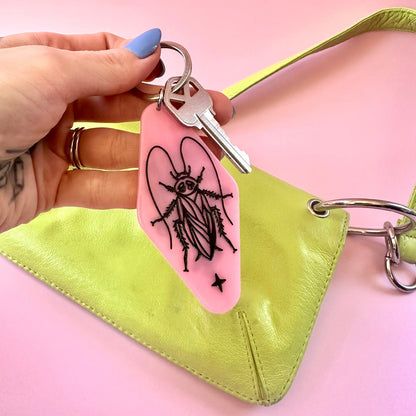 A hand with blue nail polish holds a pink keychain featuring a black insect illustration and a silver key, positioned in front of a lime green leather clutch bag on a solid pink background.