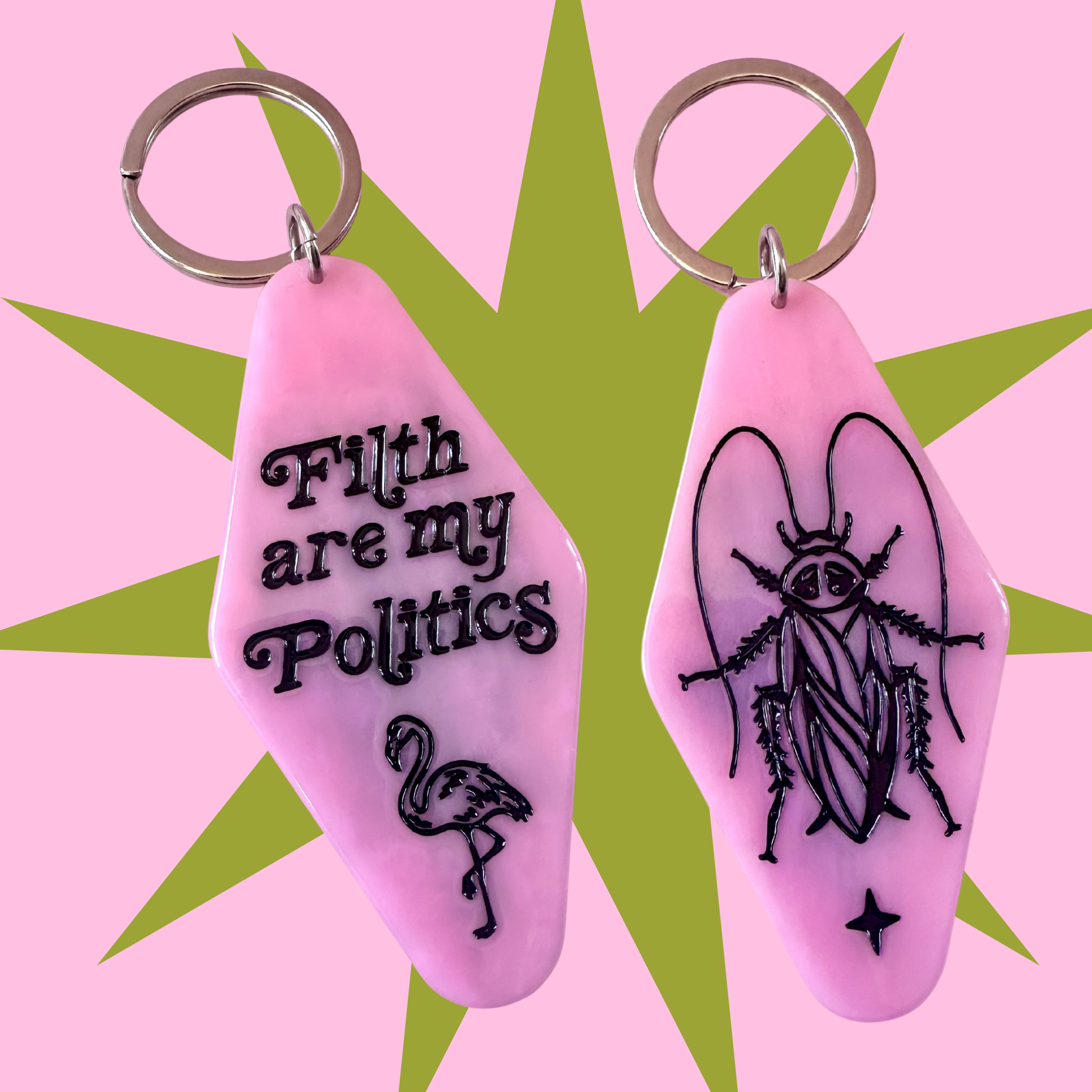 Two pink coffin-shaped keychains with silver metal key rings, displayed against a pink and green starburst background. One keychain features the black text 'Filth are my Politics' and a flamingo illustration, while the other features a black cockroach illustration.