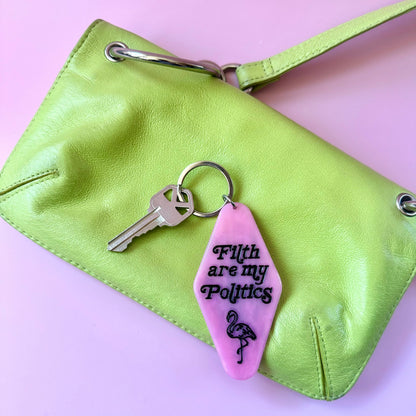 A lime green leather clutch purse with silver-tone hardware, shown on a light pink background. Attached to the keyring on the purse is a pink hotel keychain with black text reading 'Filth are my Politics' and a black flamingo graphic.