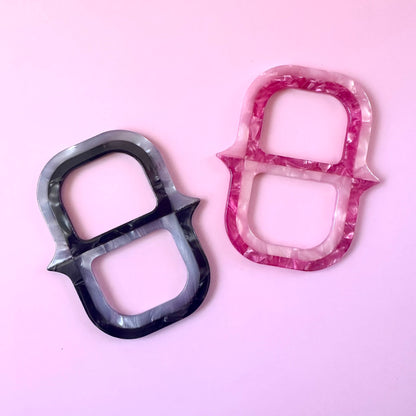 Two small chain link shaped scarf ring with a marbled pattern, one in black and gray and one in pink and white, displayed on a solid light pink background.