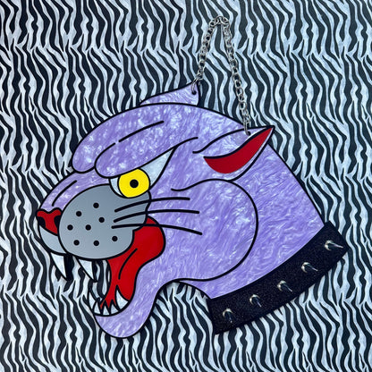 A purple panther head handbag with a textured finish, featuring a yellow eye, red accents, and a black collar with silver studs, hanging from a silver chain against a black and white zebra-print background.