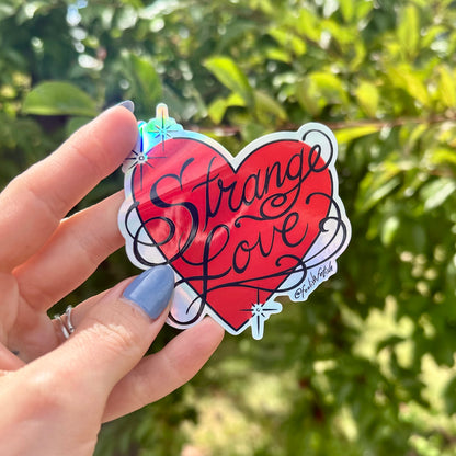 A hand with blue nail polish holds a red heart-shaped sticker that says 'Strange Love' in black script, with a holographic rainbow sheen and a white border, against a blurred green outdoor background.
